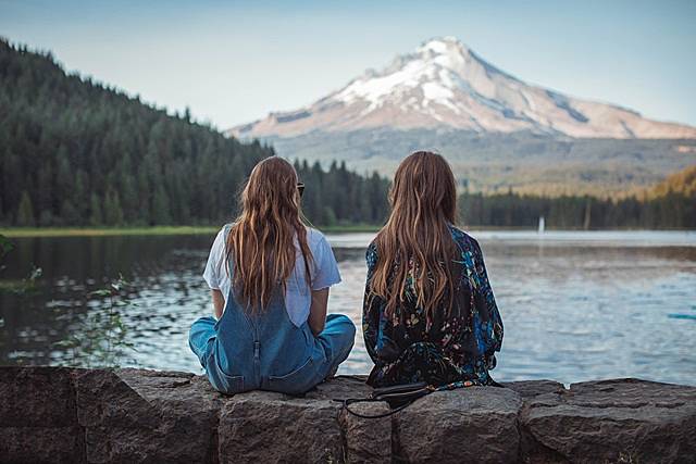 山を望む湖のほとりで座る2人の金髪の女性の後姿の画像